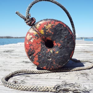 Puede incluir: Una boya redonda y desgastada, pintada en rojo y negro, con un agujero central. Una gruesa cuerda gris está atada, formando un bucle sobre la boya. La boya descansa sobre un muelle de madera, con un cielo azul claro y agua al fondo.
