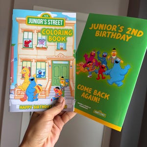 May include: Two children's books are held up. The first book is a coloring book with the text "Junior's Street Coloring Book" and features Sesame Street characters. The second book is green and says "Junior's 2nd Birthday" and "Come Back Again!"