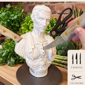 Puede incluir: Un soporte para cuchillos César blanco con diseño de busto, con tres cuchillos y unas tijeras. El texto "CAESAR KNIFE HOLDER" está en la parte superior. El soporte está sobre una base redonda gris oscuro.