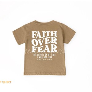 Puede incluir: Una camiseta de manga corta de color marr&oacute;n claro con el texto "FAITH OVER FEAR" en blanco. La camiseta tambi&eacute;n incluye el texto "THE LORD IS ON MY SIDE; I WILL NOT FEAR" y "PSALMS 118:6" en blanco.