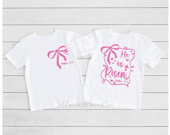 May include: Two white t-shirts with pink designs. The front of one shirt has a pink bow with hearts and the text "Matthew 28:6". The back of the other shirt has a pink bow with hearts and the text "He is Risen Matthew 28:6".