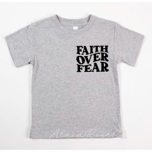 Puede incluir: Una camiseta gris de manga corta con el texto "FAITH OVER FEAR" impreso en negro en la parte delantera.