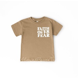 Puede incluir: Una camiseta de manga corta de color marr&oacute;n claro con las palabras "FAITH OVER FEAR" impresas en blanco en la parte delantera.