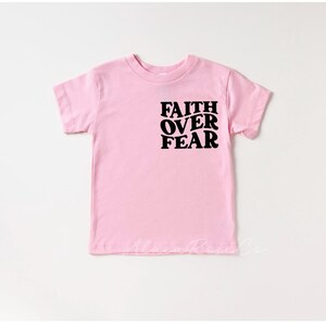 Puede incluir: Una camiseta rosa de manga corta con el texto "FAITH OVER FEAR" impreso en negro.