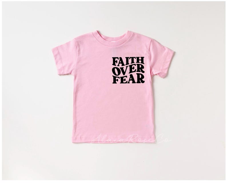 Puede incluir: Una camiseta rosa de manga corta con el texto "FAITH OVER FEAR" impreso en negro.
