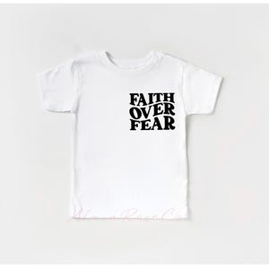 Puede incluir: Camiseta blanca con texto negro que dice "Faith Over Fear" en una fuente retro.
