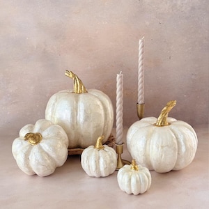 May include: A collection of white pumpkins with gold stems in various sizes, arranged with two white taper candles in gold candle holders. The pumpkins are set against a soft, neutral background, creating a fall-themed display.