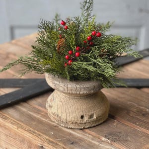 May include: A rustic wooden pedestal with a green and red holiday arrangement. The arrangement features evergreen branches, red berries, and a small pine cone. The pedestal has a weathered, aged appearance, and is set on a wooden surface.