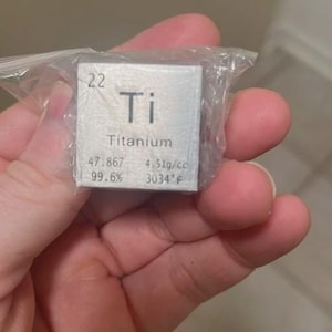 May include: A small, square titanium metal sample, encased in a clear plastic bag, is held in a hand. The sample has the element symbol "Ti" and the word "Titanium" printed on it, along with scientific data.