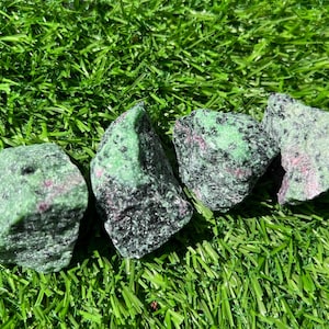 May include: Four rough-cut ruby in zoisite stones, displaying a mix of green, black, and red hues. The stones are arranged on a bed of vibrant green artificial grass, creating a natural contrast.