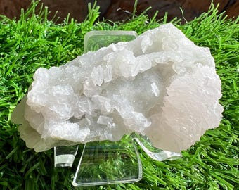 May include: A large, light-colored crystal cluster with a rough, textured surface. The crystal is displayed on a clear acrylic stand against a backdrop of green artificial grass and a brown stone wall.
