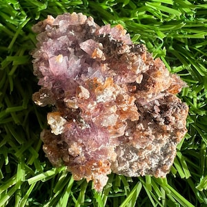 May include: A cluster of amethyst crystals with a mix of light purple and orange hues. The crystals have a rough, textured surface and are set against a backdrop of green artificial grass.