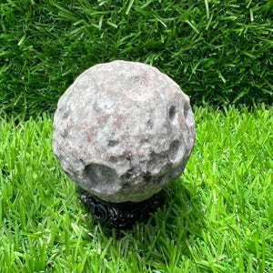 May include: A spherical stone carving with a textured, cratered surface resembling the moon, featuring mottled gray and reddish-brown mineral tones. The sphere is supported by a small, ornate black stand with intricate, carved detailing.