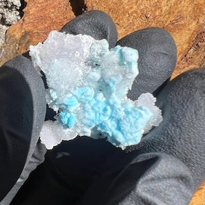 May include: A close-up of a mineral specimen featuring a cluster of light blue and white crystals. The crystals have a textured, bubbly appearance. The specimen is held in a black gloved hand, with a brown and orange rock in the background.
