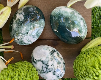 May include: Three green and white moss agate tumbled stones on a wooden surface. The stones are smooth and polished, with a variety of green and white patterns.