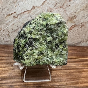 May include: A rough-hewn, natural mineral specimen with a vibrant green and black color palette. The crystalline structure is visible, and the stone is displayed on a clear acrylic stand, showcasing its raw beauty. The background features a marbled pattern.