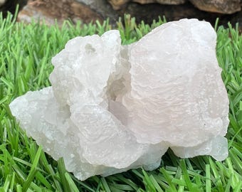 May include: A cluster of translucent, pale pink quartz crystals. The crystals have a layered, textured appearance and are resting on a bed of green grass. The background features a blurred view of rocks.