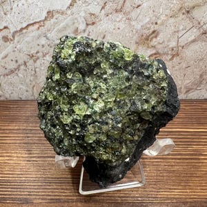 May include: A rough, dark rock formation with clusters of small, light green crystals. The rock sits on a clear display stand, showcasing its natural texture and color variations. The background features a marbled pattern.