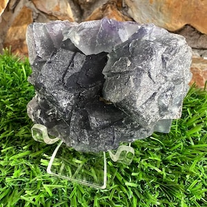 May include: A large, rough-hewn fluorite crystal in shades of gray and purple, displayed on a clear acrylic stand. The crystal's surface has a textured, crystalline appearance. The background features green grass and a stone wall.