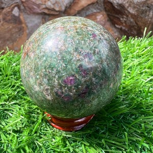 May include: A large, polished gemstone sphere with a mottled green and burgundy surface. The sphere is resting on a small, round, reddish-brown wooden stand, set against a backdrop of green artificial grass and a stone wall.