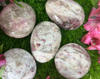 May include: Five smooth, polished, oval-shaped stones with a white and pink marbled pattern. The stones are arranged on a green surface with pink flowers.
