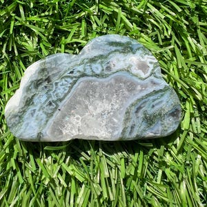 May include: A polished, cloud-shaped stone with a mix of gray, white, and blue hues, accented by green veining. The stone is set against a backdrop of vibrant green artificial grass.