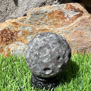 May include: A spherical stone sculpture with a textured, cratered surface in shades of dark gray and charcoal. The object is mounted on a small, ornate black circular stand, showcasing a detailed, uneven surface that resembles the moon's topography.