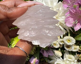 May include: A pale pink, translucent rose quartz crystal formation with a layered, jagged, and crystalline structure. The mineral specimen displays a soft, semi-transparent appearance with sharp, angular edges and a natural, unpolished surface texture.