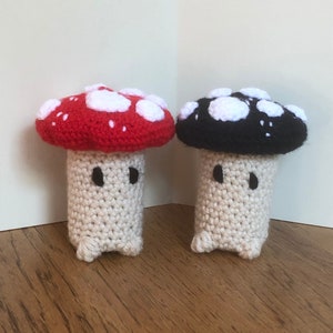 Pode incluir: Duas figuras de cogumelos de crochê. Um cogumelo tem uma tampa vermelha com manchas brancas, o outro uma tampa preta com manchas brancas. Ambos têm caules esbranquiçados com pequenos olhos pretos e pés minúsculos. Os cogumelos são feitos à mão.
