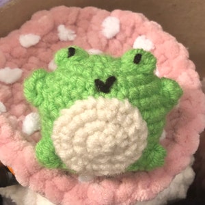 Pode incluir: Um sapo de crochê feito à mão em verde brilhante, com barriga branca e olhos e boca pretos. O sapo está sentado em cima de uma almofada rosa em forma de cogumelo com manchas brancas. O brinquedo de pelúcia é macio e texturizado.