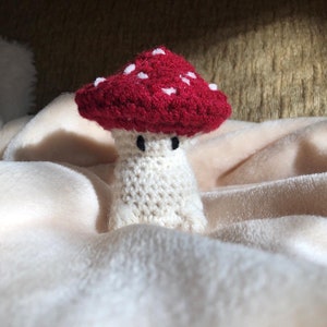 Pode incluir: Um brinquedo de cogumelo de crochê com um chapéu vermelho e branco e um corpo branco. O cogumelo tem olhos pretos e está sentado em um cobertor branco.