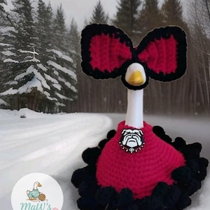 May include: A white goose figurine wearing a red and black crocheted outfit. The outfit includes a red dress with a black ruffled hem and a matching red and black bow on its head. A bulldog emblem is attached to the center of the red dress.