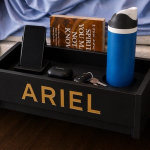 May include: A black rectangular bedside caddy with the name ARIEL in gold lettering on the front. The caddy holds a black smartphone, a black earbud case, a set of keys, a blue water bottle, and a book titled Spirit You May Not Know.