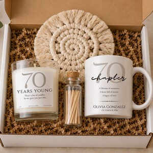 May include: A gift box containing a glass candle with the text 70 YEARS YOUNG, a round cream macrame coaster, a small glass bottle filled with wooden matches, and a white ceramic mug featuring the text 70 chapter, Olivia Gonzalez, and October 8, 1956.