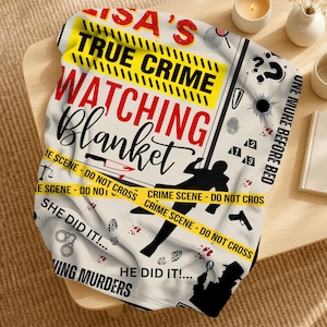 May include: A cream-coloured blanket with a crime scene theme. The blanket features the text "LISA'S TRUE CRIME WATCHING Blanket" in red and black, along with crime scene tape, silhouettes, and other crime-related graphics.