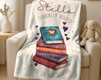 May include: A cream-coloured throw blanket draped over a chair, featuring a stack of illustrated books with heart and star accents. The blanket has the text "Stella Currently Booked Please Do Not Disturb."