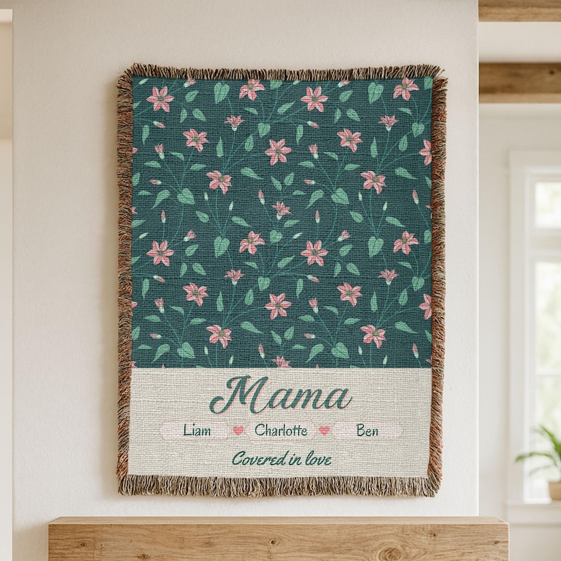 Pu&ograve; includere: Una coperta tessuta con un motivo floreale verde acqua e bordi sfrangiati. La coperta presenta la parola "Mama" in corsivo, con nomi e la frase "Covered in love" sotto.