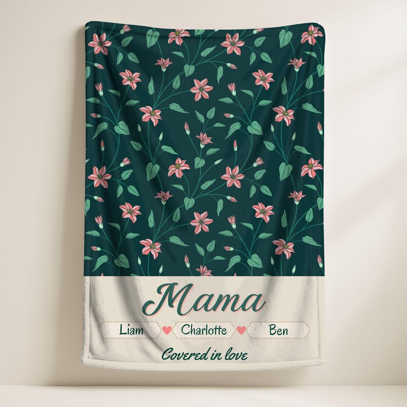 Pu&ograve; includere: Una coperta verde scuro con un motivo floreale di fiori rosa e foglie verdi. La sezione inferiore presenta la parola "Mama" in corsivo, con nomi e cuori, e la frase "Covered in love".