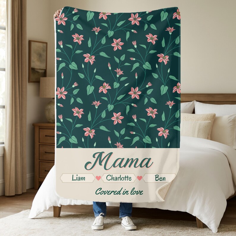Pu&ograve; includere: Una coperta verde scuro con un motivo floreale di fiori rosa e foglie verdi. La sezione inferiore presenta la parola "Mama" in corsivo, con nomi e cuori sottostanti. &Egrave; presente anche la frase "Covered in love".
