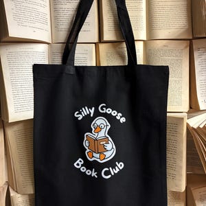 Può includere: Borsa tote nera con la scritta "Silly Goose Book Club" e un'oca dei cartoni animati che legge un libro. La borsa ha due manici neri ed è ambientata su uno sfondo di libri aperti.
