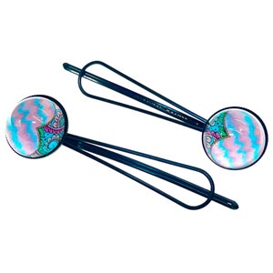 May include: Two dark blue bobby pins with round, decorative tops. The tops feature a colorful design with pink, blue, and green patterns. The bobby pins are set against a white background.