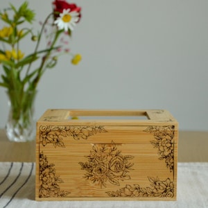 Könnte beinhalten: Rechteckige Bambusbox mit durchsichtigem Deckel und Blumengravuren. Die Box ist hellbraun und hat ein detailliertes Blumendesign auf der Vorderseite und an den Ecken. Im Hintergrund eine Vase mit Blumen.