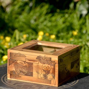 Op de afbeelding: Een houten doos met een glazen deksel, met een Japans geïnspireerd ontwerp met een kat die een munt vasthoudt en een wolkpatroon. De doos is gemaakt van lichtbruin hout en heeft een natuurlijke afwerking.