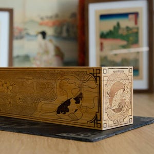 May include: A rectangular wooden box featuring intricate laser-engraved designs on its side and end panels. The side panel displays a koi fish swimming among cherry blossoms and wave patterns, while the end panel depicts a fan and circular motif.