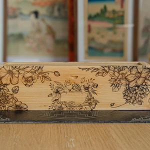 Pode incluir: Uma caixa retangular de madeira clara com um padrão floral detalhado gravado a laser em castanho escuro no painel frontal. O design inclui várias flores, folhas e caules dispostos numa ilustração botânica contínua.