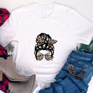 Op de afbeelding: Wit T-shirt met een afbeelding van een vrouw met een rommelige knot, een haarband met luipaardprint en een zonnebril. De afbeelding is in zwart, bruin en wit.