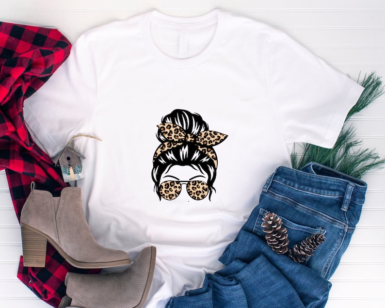 Op de afbeelding: Wit T-shirt met een afbeelding van een vrouw met een rommelige knot, een haarband met luipaardprint en een zonnebril. De afbeelding is in zwart, bruin en wit.