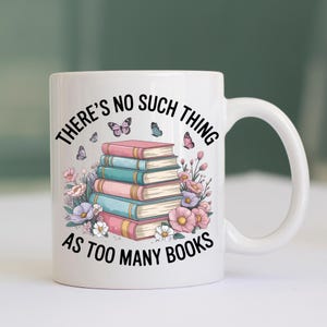 以下が含まれることがあります： 「THERE'S NO SUCH THING AS TOO MANY BOOKS」の文字が入った白いセラミックマグ。ピンクとティール色の本が積み重ねられ、蝶と花が描かれています。