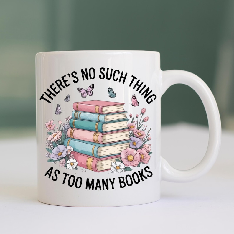 以下が含まれることがあります： 「THERE'S NO SUCH THING AS TOO MANY BOOKS」の文字が入った白いセラミックマグ。ピンクとティール色の本が積み重ねられ、蝶と花が描かれています。