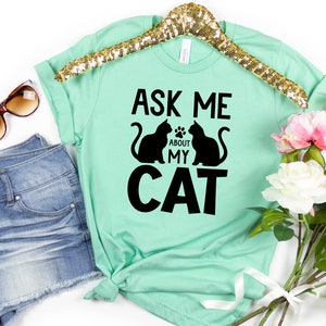 Może przedstawiać: Mint zielony t-shirt z czarnym tekstem, kt&oacute;ry brzmi "Ask me about my cat" i dwie czarne sylwetki kot&oacute;w.